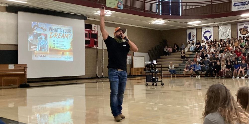 man in black shirt talking to the gymnasium full of students. His hand is in the air with a microphone in the other hand.