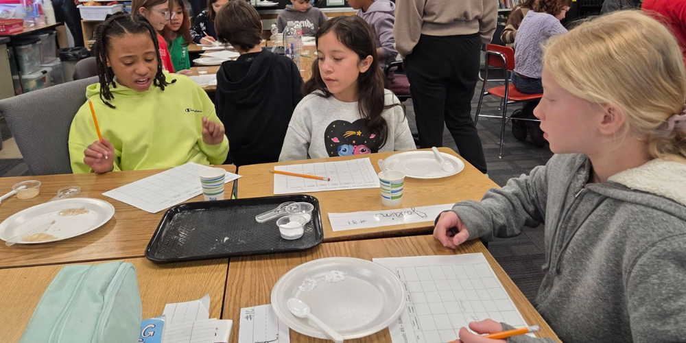Three girls are seated around a group of desks, filling out a lab paper while experimenting with baking ingredients. 