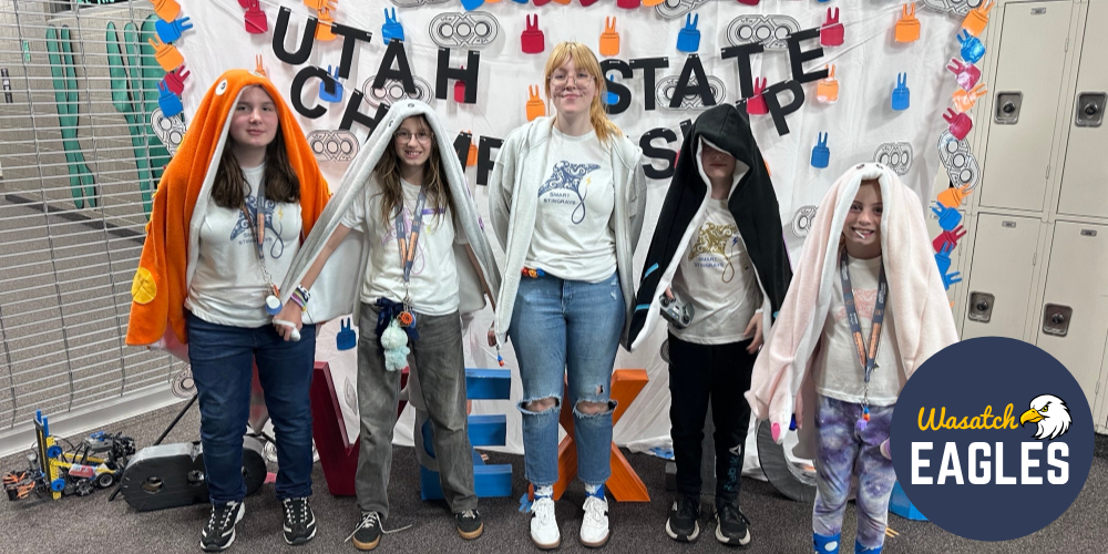 Group of elementary students standing indoors in front of a large handmade display that reads “Utah State University.” The backdrop is decorated with colorful paper handprints and gear-themed cutouts, with robotics pieces and cables visible on the floor nearby. The students are wearing casual clothing, including jeans, sneakers, and matching white shirts, and several have light jackets or blankets draped over their heads and shoulders. The setting appears to be a school or campus hallway with lockers, tile flooring, and a metal gate in the background.