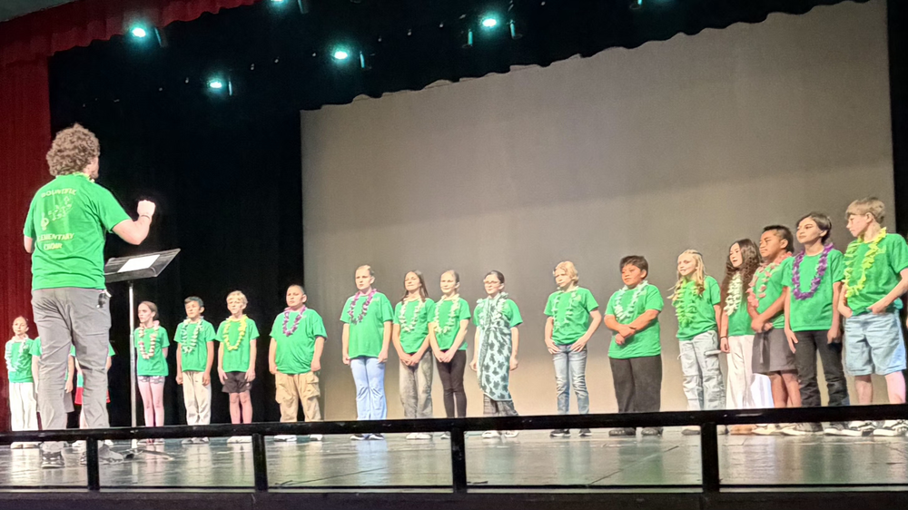 Bountiful Elementary choir students perform on stage during a school music event, wearing green shirts and leis while their director leads from the front.