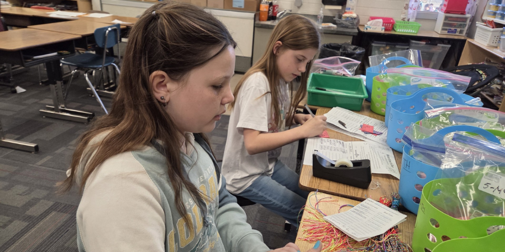 Two girls sitting at desks with basket on them. They are filling out tags for the friendshipgrams and taping a friendship bracelet on them. 