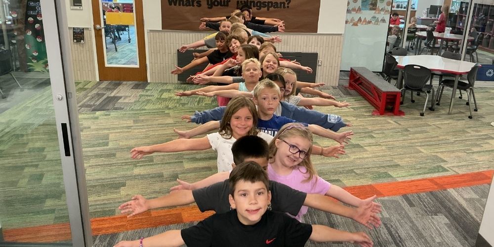A class of students face the camera in a line to the far wall, arms outstretched under a “What’s Your Wingspan?” sign.