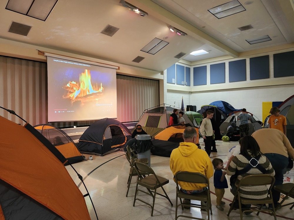 Tents set up in the gym with a fire projected on the screen. Kids and teachers standing and sitting on chairs.
