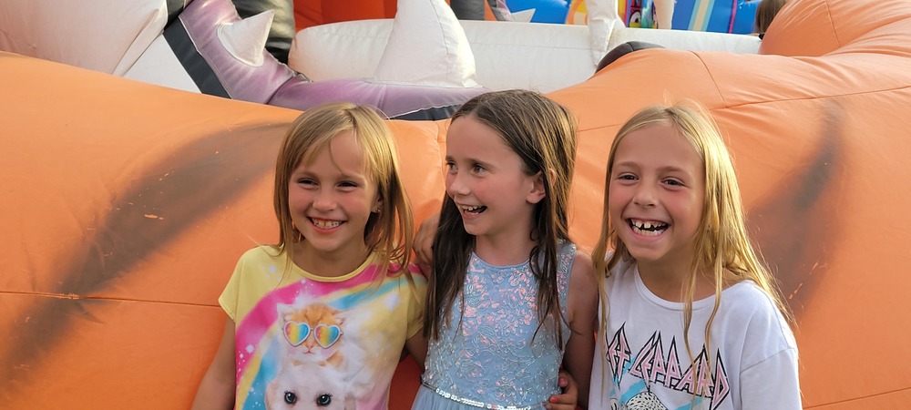 three girls laughing at the school carnival in front of a tiger blow-up slide.