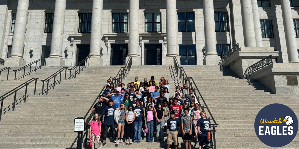 Fourth grade students from Wasatch Elementary pose for a group photo on the grand front steps of the Utah State Capitol. The large stone building with tall columns rises behind the students, who are gathered in the center of the staircase. Black handrails divide the wide steps, and bright sunlight highlights the Capitol's historic architecture. A Wasatch Eagles logo appears in the bottom-right corner of the image.