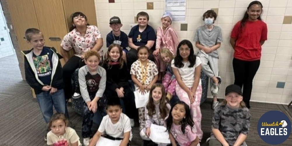 Group photo of approximately 18 Wasatch Elementary students recognized as September Students of the Month. The students are gathered in three rows inside a school hallway with tiled walls and a wooden door in the background. Several students are holding certificates while others are seated or standing around them. The students are wearing a variety of school and pajama-themed clothing. The Wasatch Eagles logo appears in the lower-right corner of the image.
