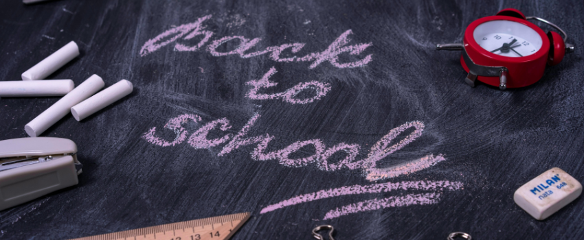 Back to school written in pink chalk on a blackboard, surrounded by school supplies including white chalk sticks, a red alarm clock, a stapler, a ruler, an eraser, and binder clips.