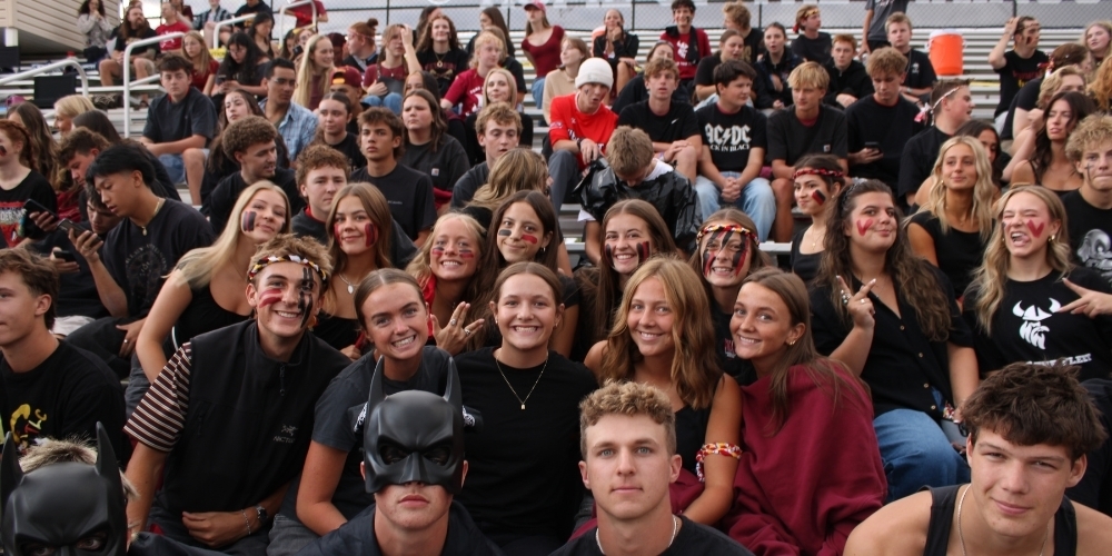Large group of Viewmont High students packed on bleachers, many in black/red with face paint and headbands, smiling and throwing peace signs; two front students in Batman masks.
