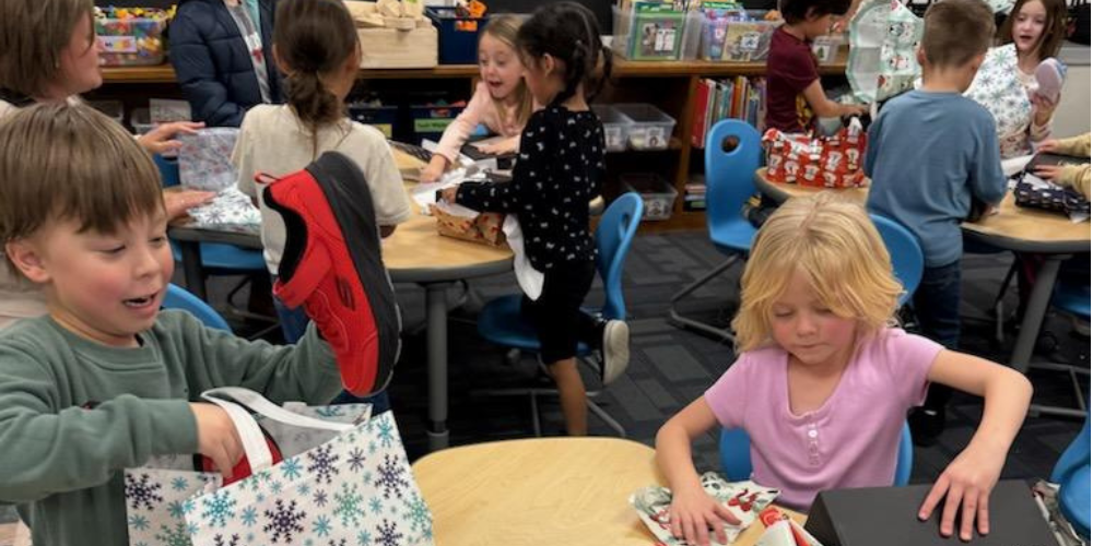 Kindergarten boys and girls  sitting at tables in a classroom opening wrapped boxes of shoes.. 