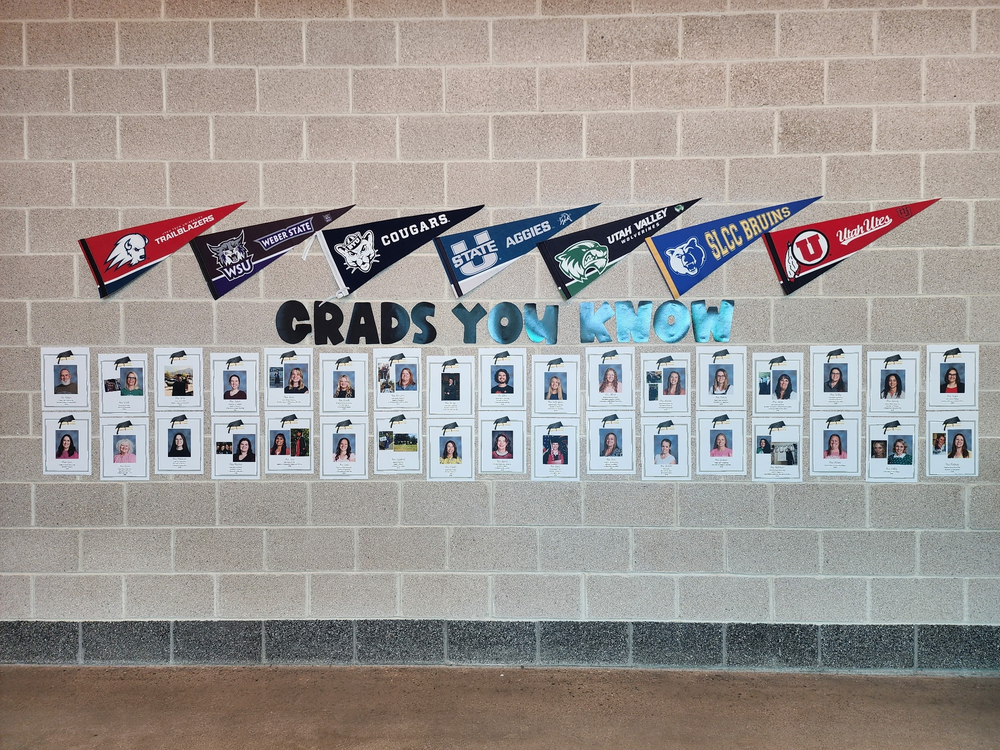 Wall display at Bountiful Elementary featuring the phrase “Grads You Know” in large letters, with college pennants across the top and staff graduation photos arranged in rows below on a brick wall.