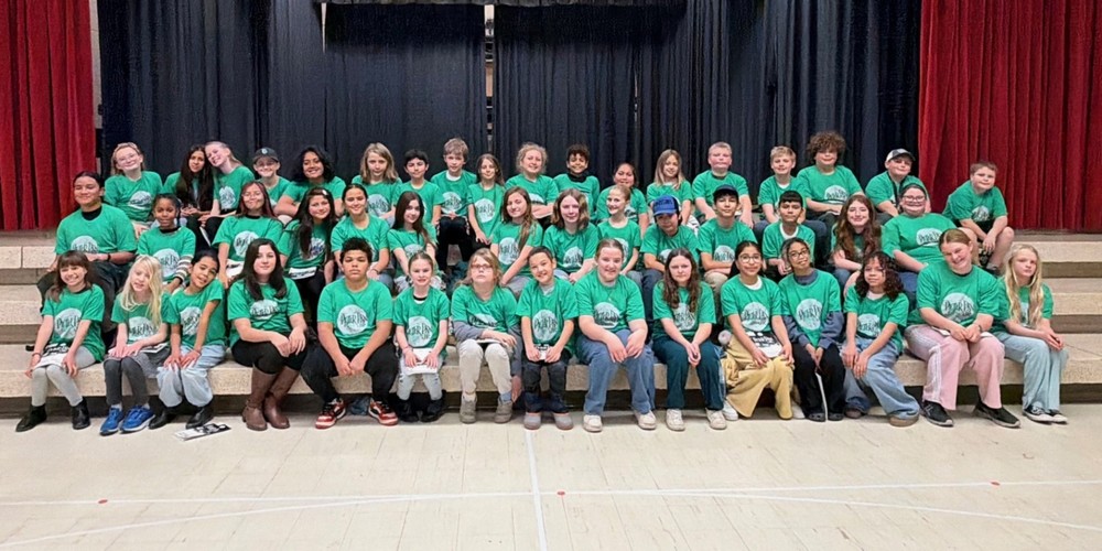 The cast of Peter Pan Jr on the steps on the stage. All wearing a green cast shirt that says Peter Pan Jr. The steps are light in color, the back curtains are black, the front curtains are red