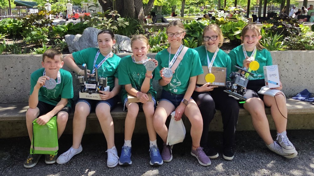 Six Bountiful Elementary students sit together outdoors at the Utah Elementary Robotics competition at Lagoon, smiling in green team shirts and wearing UER medals while showing their robots, trophies, and awards from the competition.