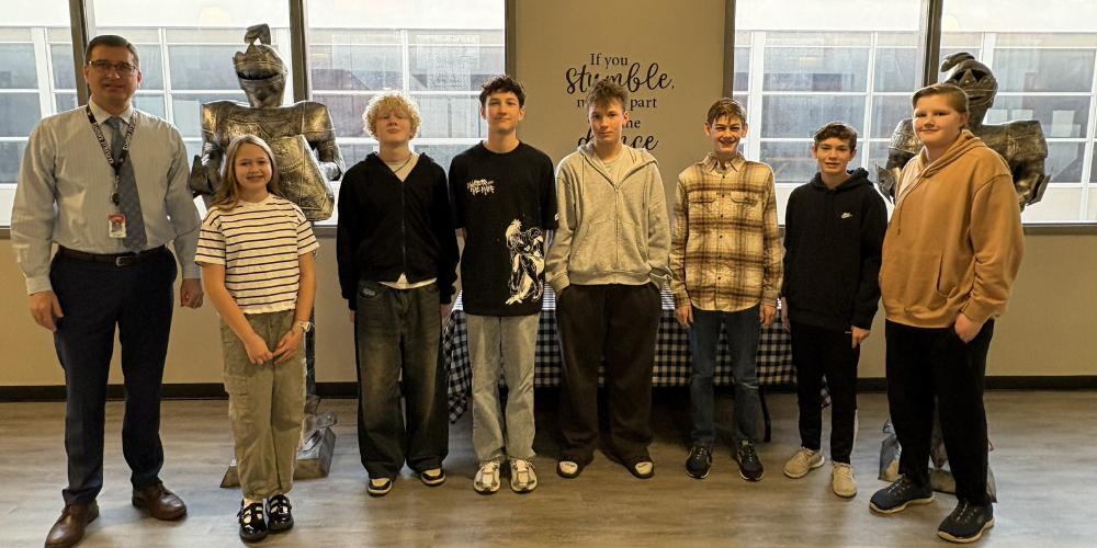 image of 7 students and the principal standing in front of two knight statues and a table.