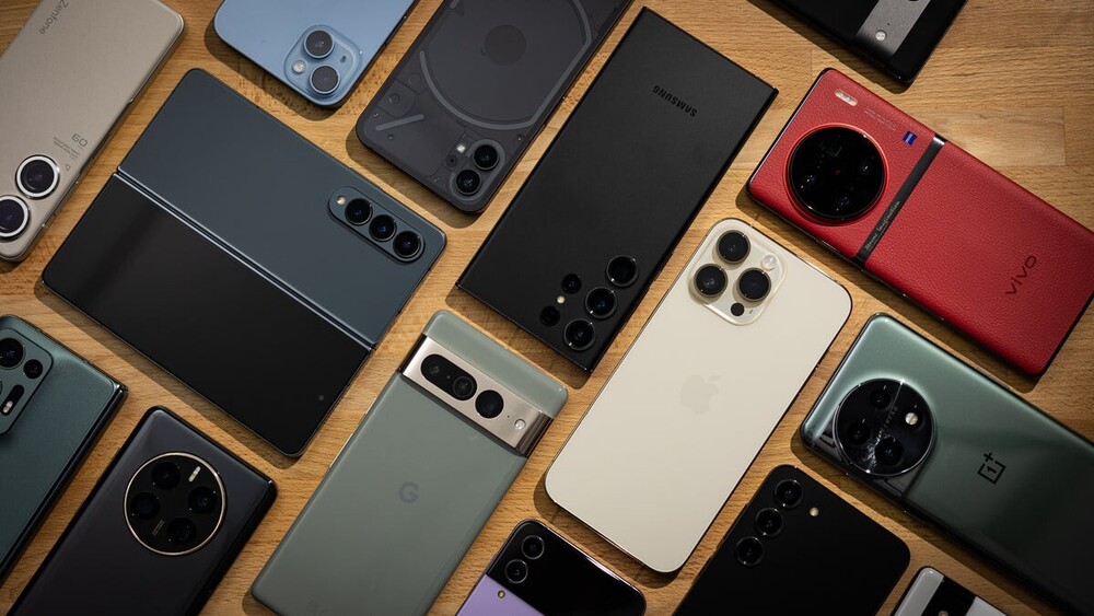 Several smartphones from different manufacturers are arranged on a wooden surface. The devices vary in size, color, and design, including both standard and foldable models, with their backs and camera arrays visible. The image represents a variety of personal electronic devices.