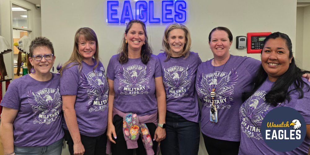 A group of six adults stand indoors beneath a blue “EAGLES” sign on a school wall. Each person is wearing a purple T‑shirt with the words “Purple Up for Military Kids.” The group is positioned shoulder to shoulder in a school hallway, with doors and wall fixtures visible in the background. A “Wasatch Eagles” logo appears in the lower right corner of the image.