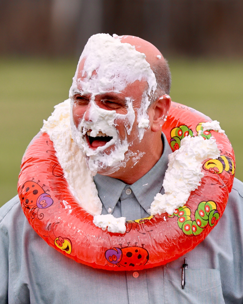 Principal Darryl Denhalter with  smashes whipped cream all over his face. he is smiling and wearing a floaty around his neck.