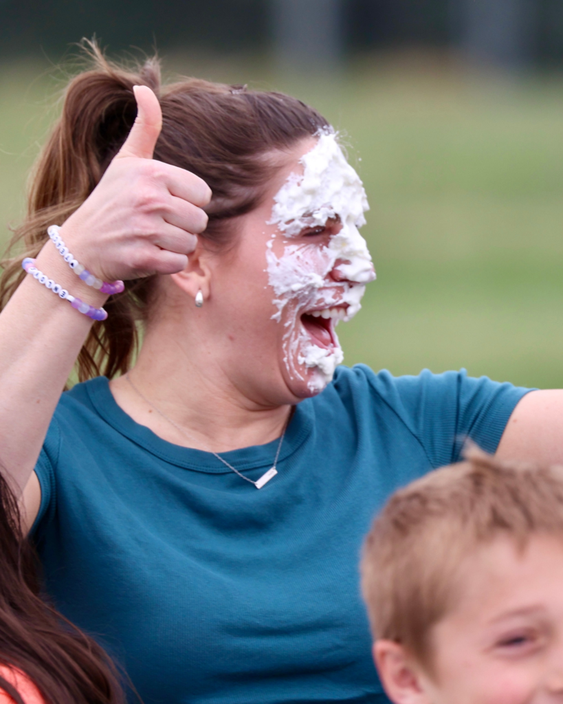 A teacher smiles and gives a thumbs up after she has a pie shoved in her face.