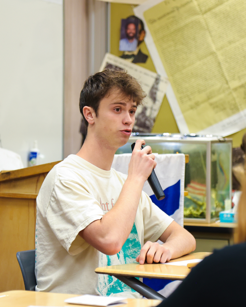 A student sitting at a desk holding a microphone speaks.