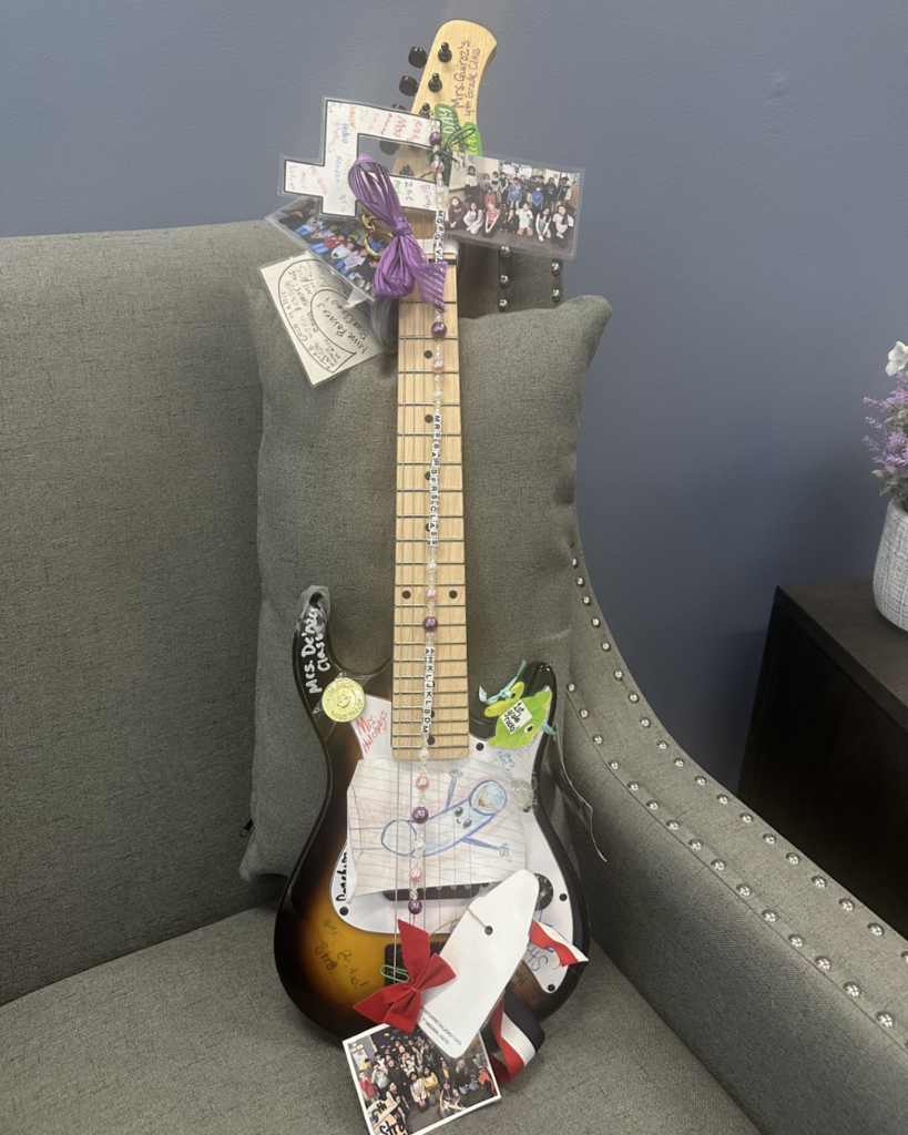 A photo of a guitar sitting on a chair. The guitar has drawings, bows, and other things tied to it.