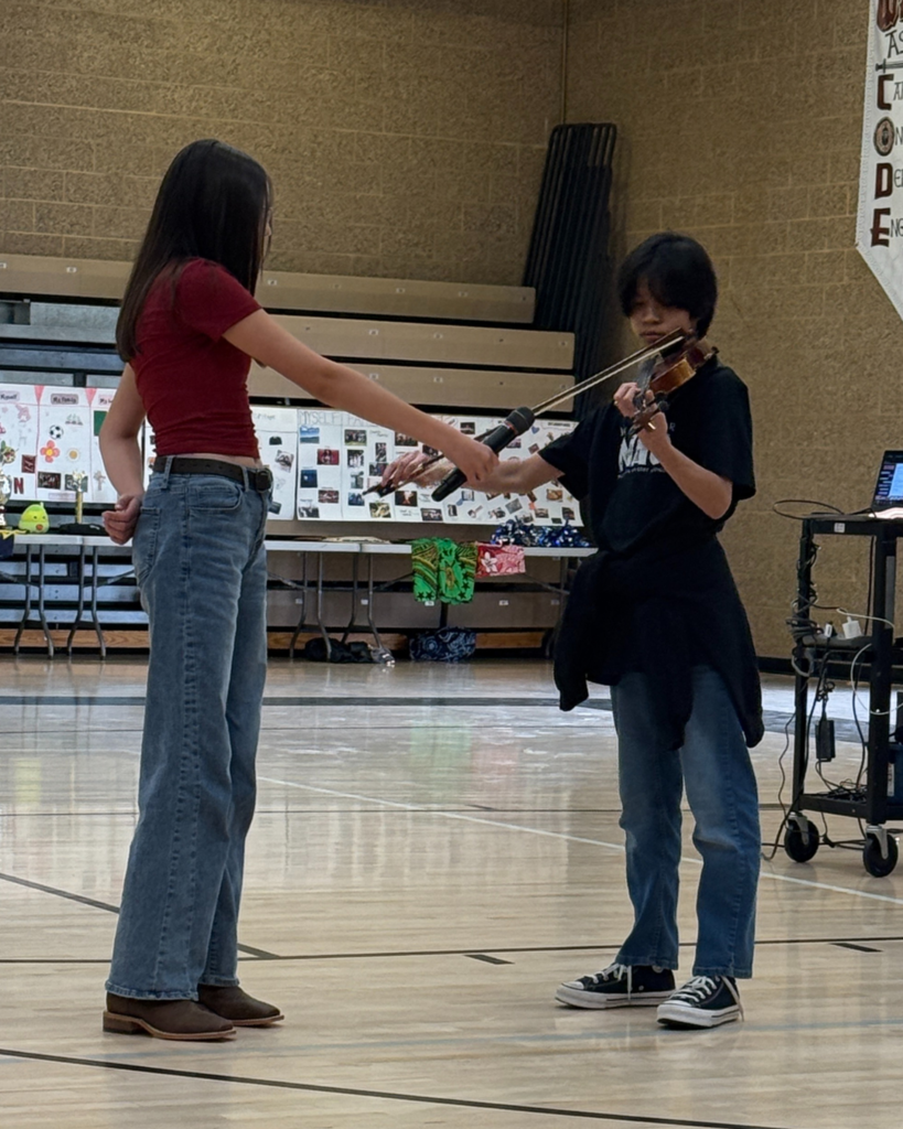 A student holds a microphone up to another student playing a string instrument, probably a violin but may be a viola.