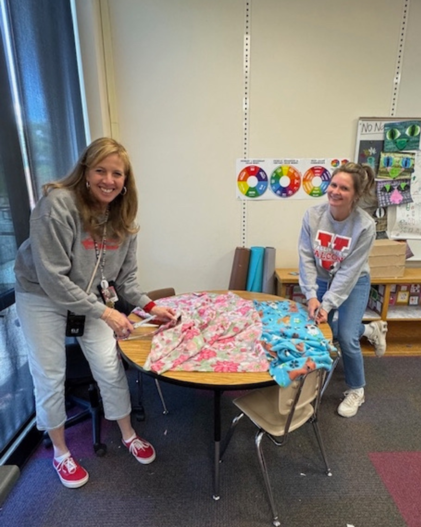 Two faculty members at Valley View Elementary smile at the camera as they tie blankets at school on their desks.