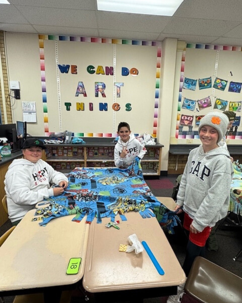 Three students smile at the camera as they tie blankets at school on their desks.
