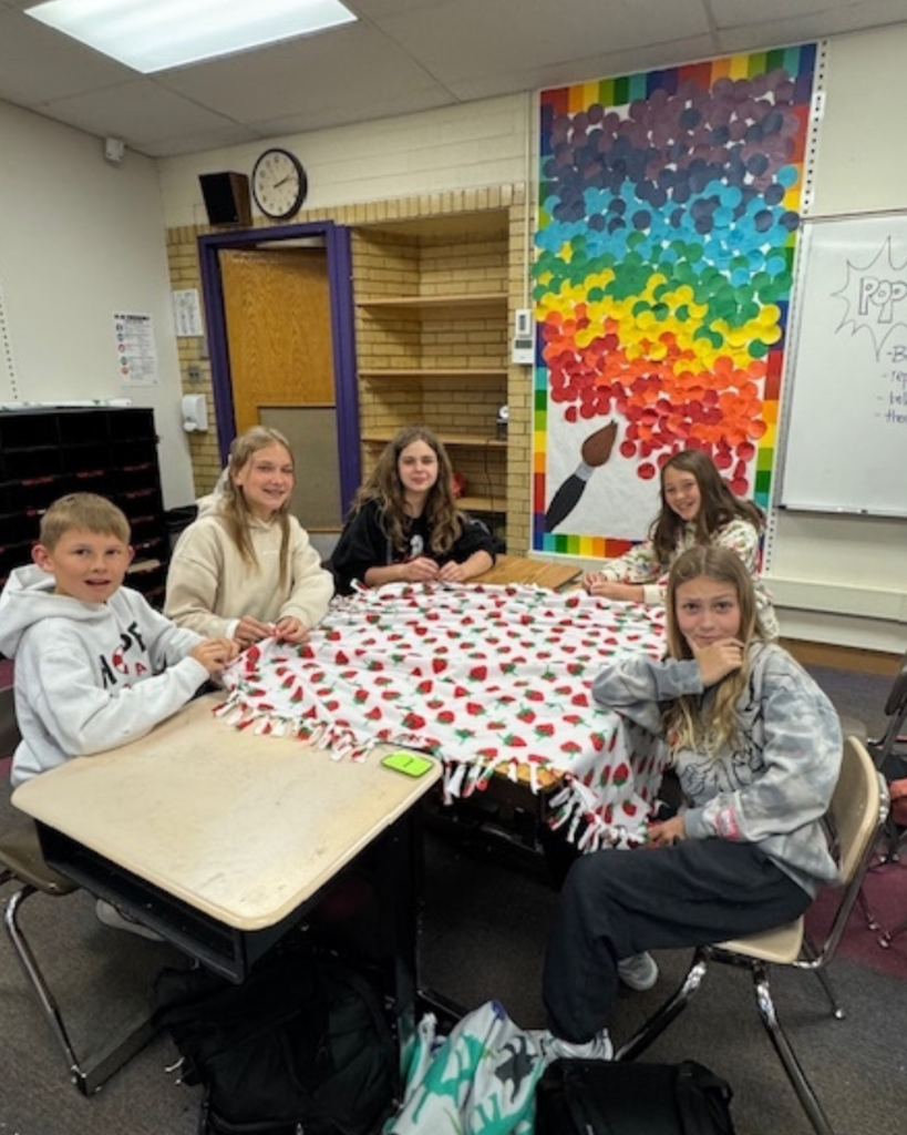 Five students smile at the camera as they tie blankets at school on their desks.