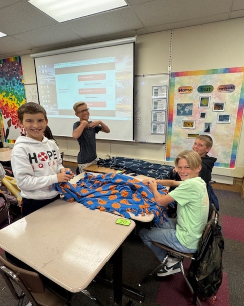 Four students smile at the camera as they tie blankets at school on their desks.
