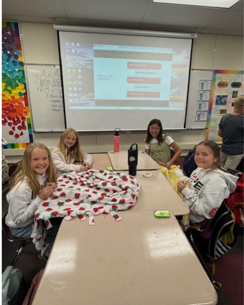 Four students smile at the camera as they tie blankets at school on their desks.