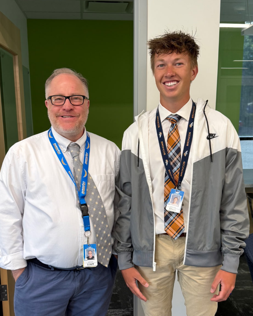 A principal stands and smiles at the camera with one of his staff members. They are dressed up similarly today.