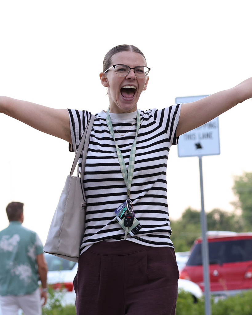A new educator smiles with a open mouth and their arms stretched wide as they walk through a crowd of people.