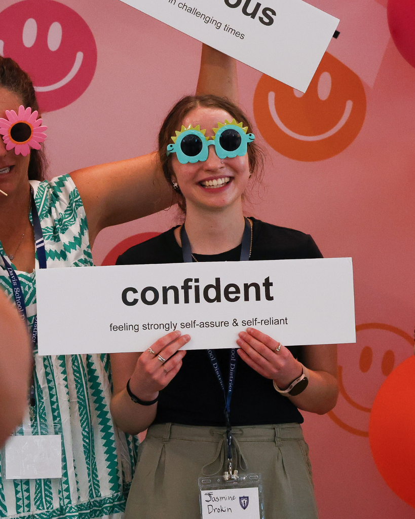 A photo of a new educator standing in front of a pink and orange smiley face backdrop. She is wearing blue and yellow dinosaur sunglasses and is holding a sign that reads: confident. Feeling strongly self-assure & self-reliant.