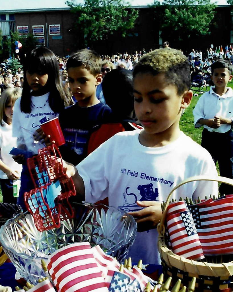 A student puts a piece of paper in a bowl that is surrounded by flags. The student is wearing a white shirt that says Hill Field Elementary.