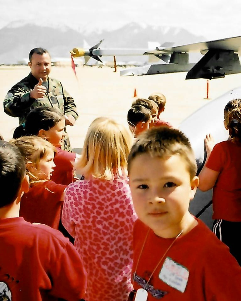 An Air Force person gives a thumbs up to a large crowd of students dressed in red. One student is looking directly at the camera. It looks like they are on a runway.