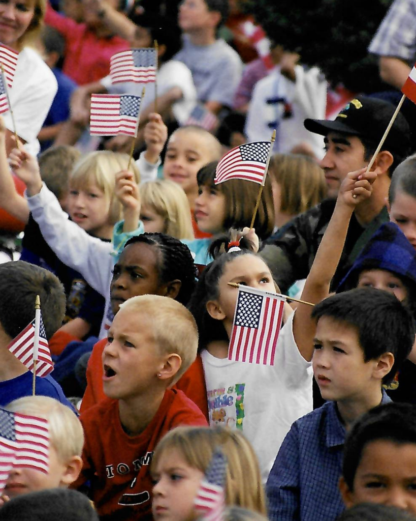 An image of children and military personnel sitting down and waving small United States flags.