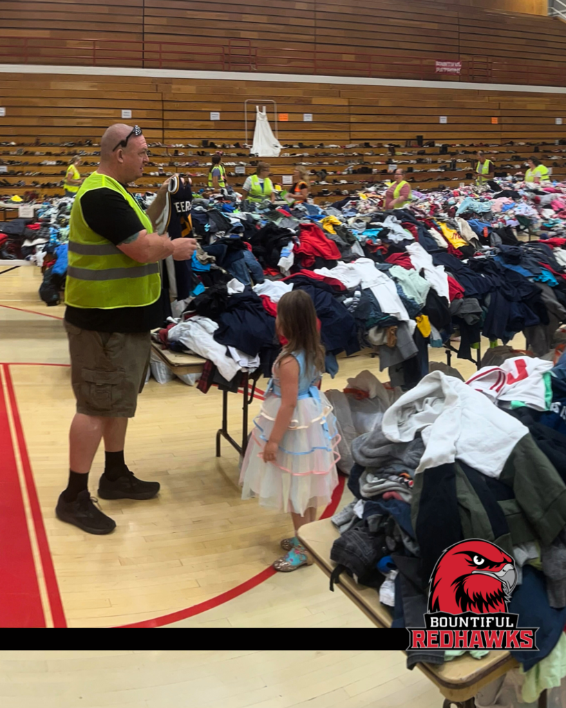 An image of many tables piled high with clothing. There are people in yellow safety vests. There is the Bountiful High logo in the bottom right corner.