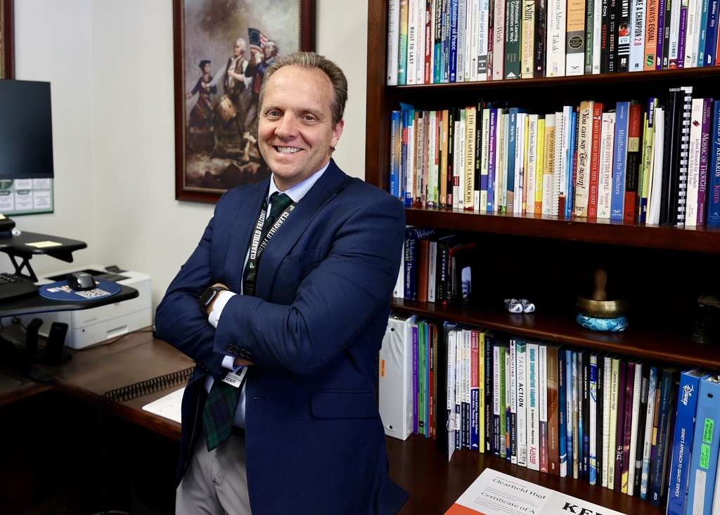 A principal folds his arms and smiles while in his office.