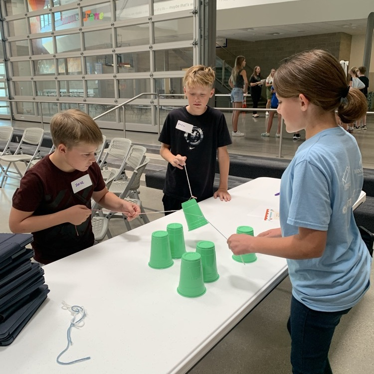 three kids work together to pick up cup using rubber bands
