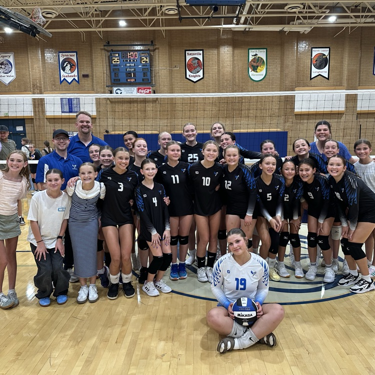 The Fairfield Falcons volleyball team standing in 2 or 3 rows. The coaches, players, and managers are all smiling. One girl is sitting in front, holding a volleyball. Behind the team is a net and the scoreboard showing our winning score of 25-24 from the last game we played.