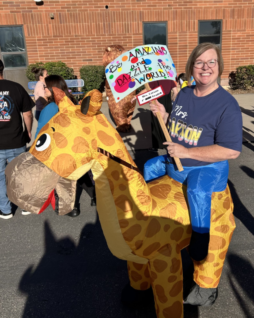 An educator wearing a blow up giraffe costume is holding a sign that says Be amazing and dazzle the world.