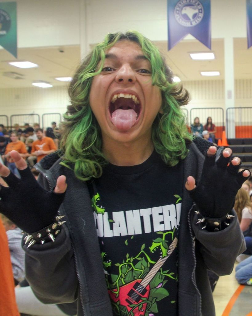 A student with green hair stands with his mouth open making a funny face and hands up like claws. 