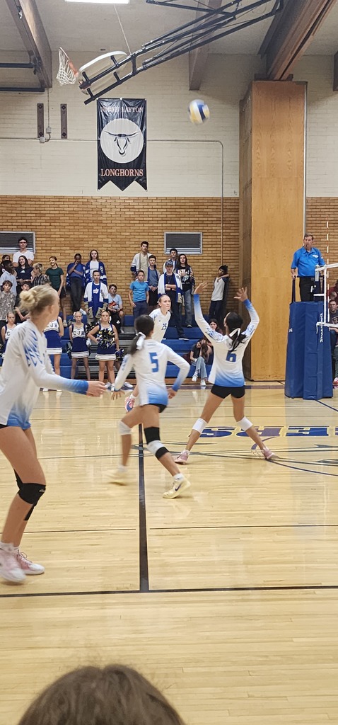 Four Fairfield players are preparing to attack a ball. The setter is receiving the ball on the right side of the pic, with the three front row hitters lining up to attack the ball at the net. In the background are the Centerville students standing in the bleachers, and a ref on a ref-stand to the right of the picture.