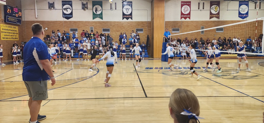 Fairfield's libero, wearing a black jersey, is passing the ball towards the right side of the picture. The setter, wearing white is running to get to the pass. The three girls from the front row are moving towards a hitting position. In the background are Centerville people in the bleachers, and the ref standing in the ref-stand.