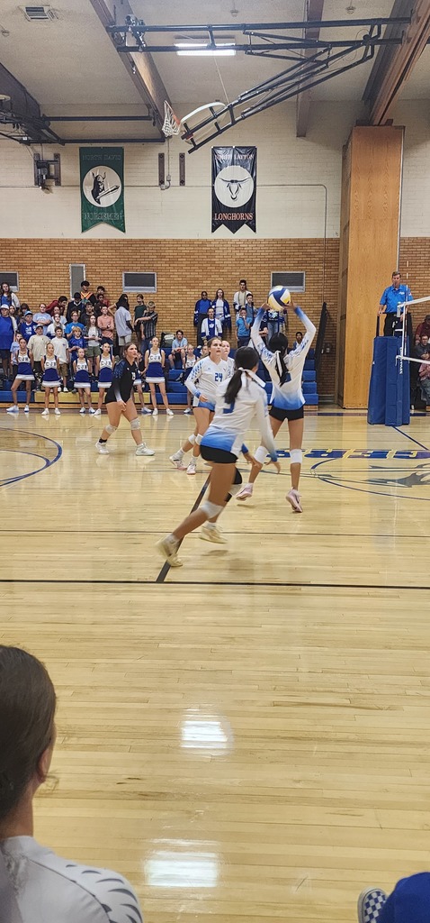 Fairfield setting the ball with the front row players approaching to hit. Behind are Centerville people in the bleachers, with a ref standing in the ref-stand on the right side of the picture.