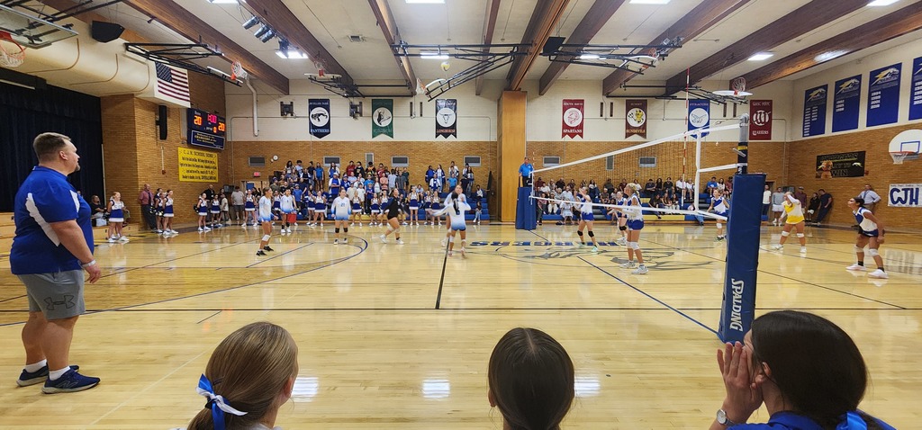 Fairfield ladies are looking up at a volleyball. The girl on the right is preparing to set the ball, the girl directly behind her is lining up to hit the ball. The three girls to the left side are watching. The background has a crowd of people in the bleachers.