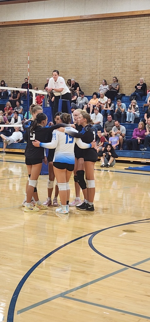 Fairfield's six volleyball players are standing in a circle, with their arms on each other's shoulders, as they talk about their game. The audience is in the bleachers to the back of the picture.