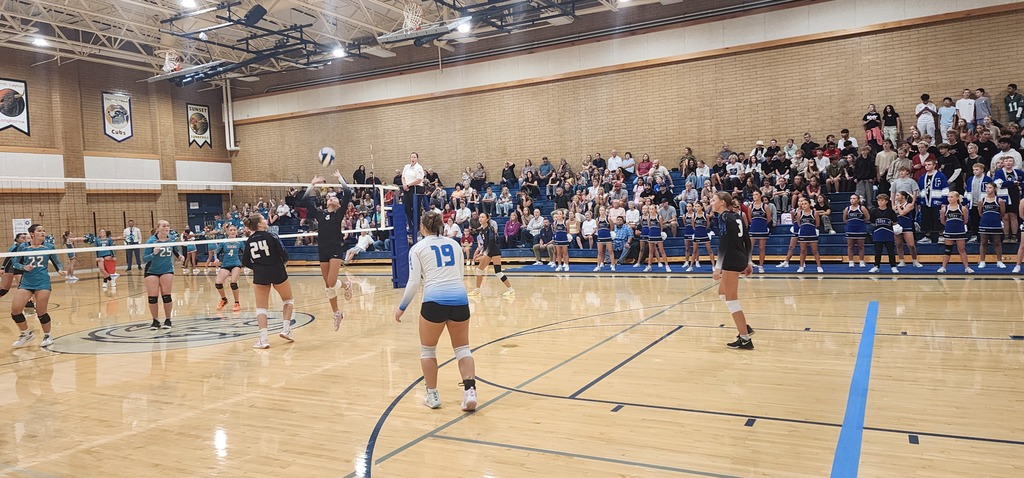 Fairfield's setter is sending a perfect set to the opposite hitter in the back of the image. Fairfield's players are watching and ready close to the front, and Horizon's players are watching on the other side of the net, towards the back of the image. In the background are fans in the bleachers.