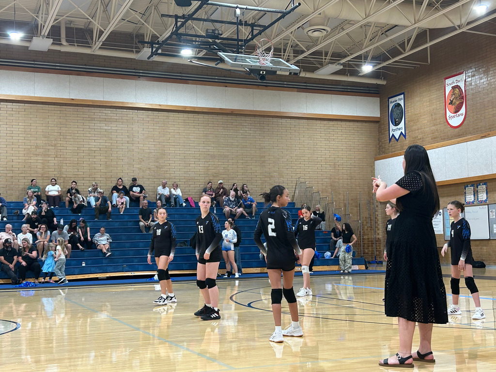 Coach Van Deusen is in the front right side of the image coaching the team. Four of Fairfield's players are spread out on the court, looking at Coach Van Deusen. In the background there are fans sitting in the bleachers.