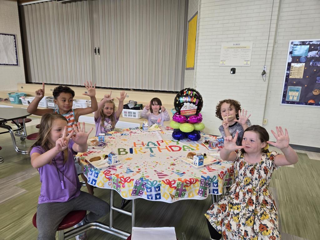 July and August Birthdays kids smiling while sitting at a round table with birthday balloons and a happy birthday table cloth