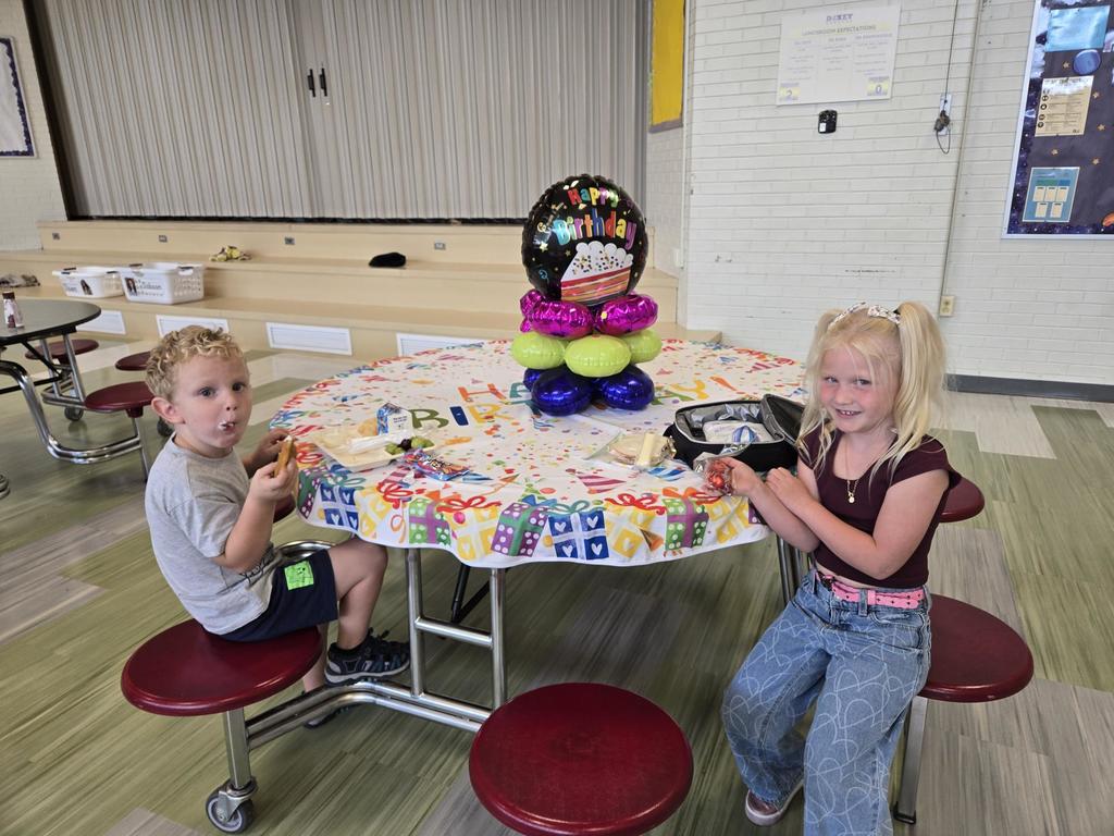 July and August Birthdays kids smiling while sitting at a round table with birthday balloons and a happy birthday table cloth